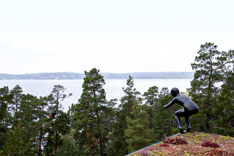 En staty av en man som står på ett tak. Vatten och träd i bakgrunden.