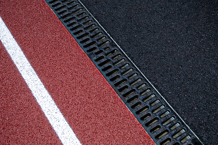 Närbild på löparbana, ränna med svart galler samt asfalt.