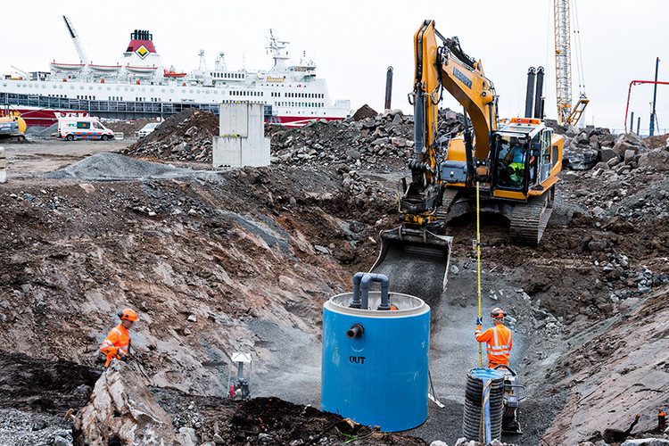 En byggarbetsplats med kranbil, blå betongtank, byggarbetare och en färja i bakgrunden.