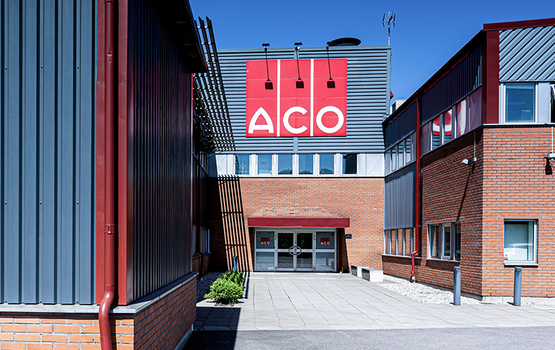 Entré med röd ACO-logotyp ovanför tegelbyggnad.